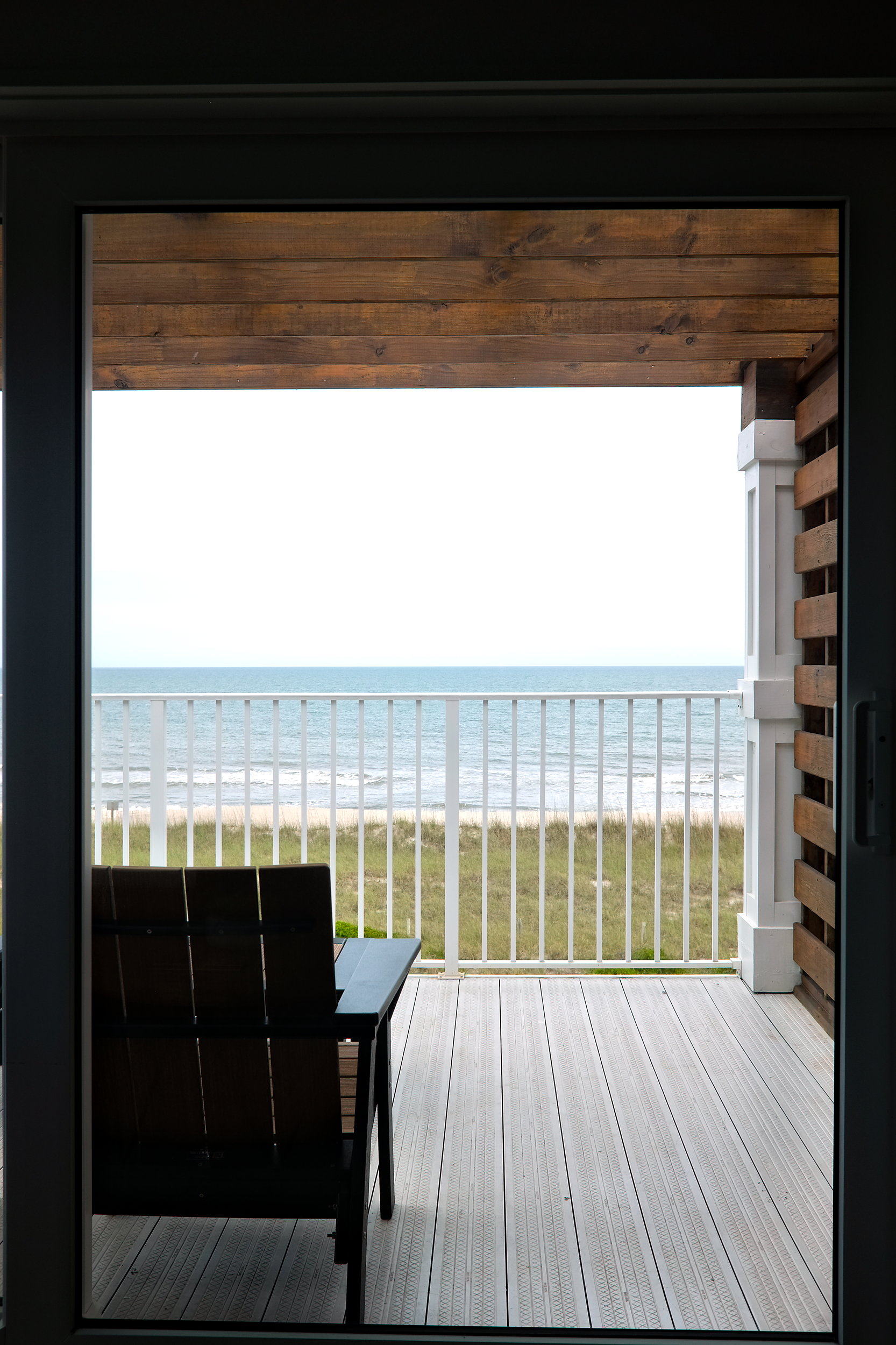 View from inside a beachfront room looking out through a glass door to a wooden balcony with a single chair and table, overlooking grassy dunes and the Atlantic Ccean under a cloudy sky.