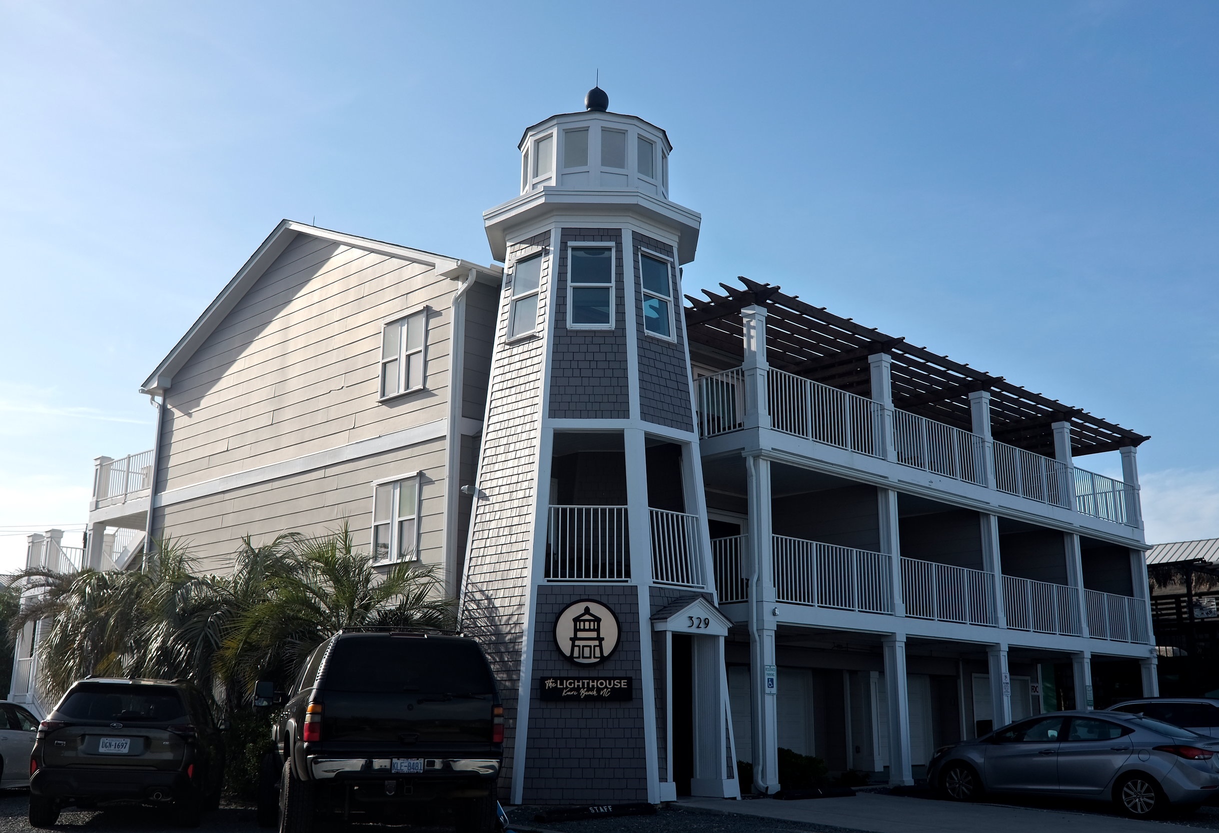 Exterior of The Lighthouse Inn in Kure Beach, North Carolina, featuring a lighthouse-style tower with signage and multiple balconies, parked cars, palm trees, and a clear blue sky.