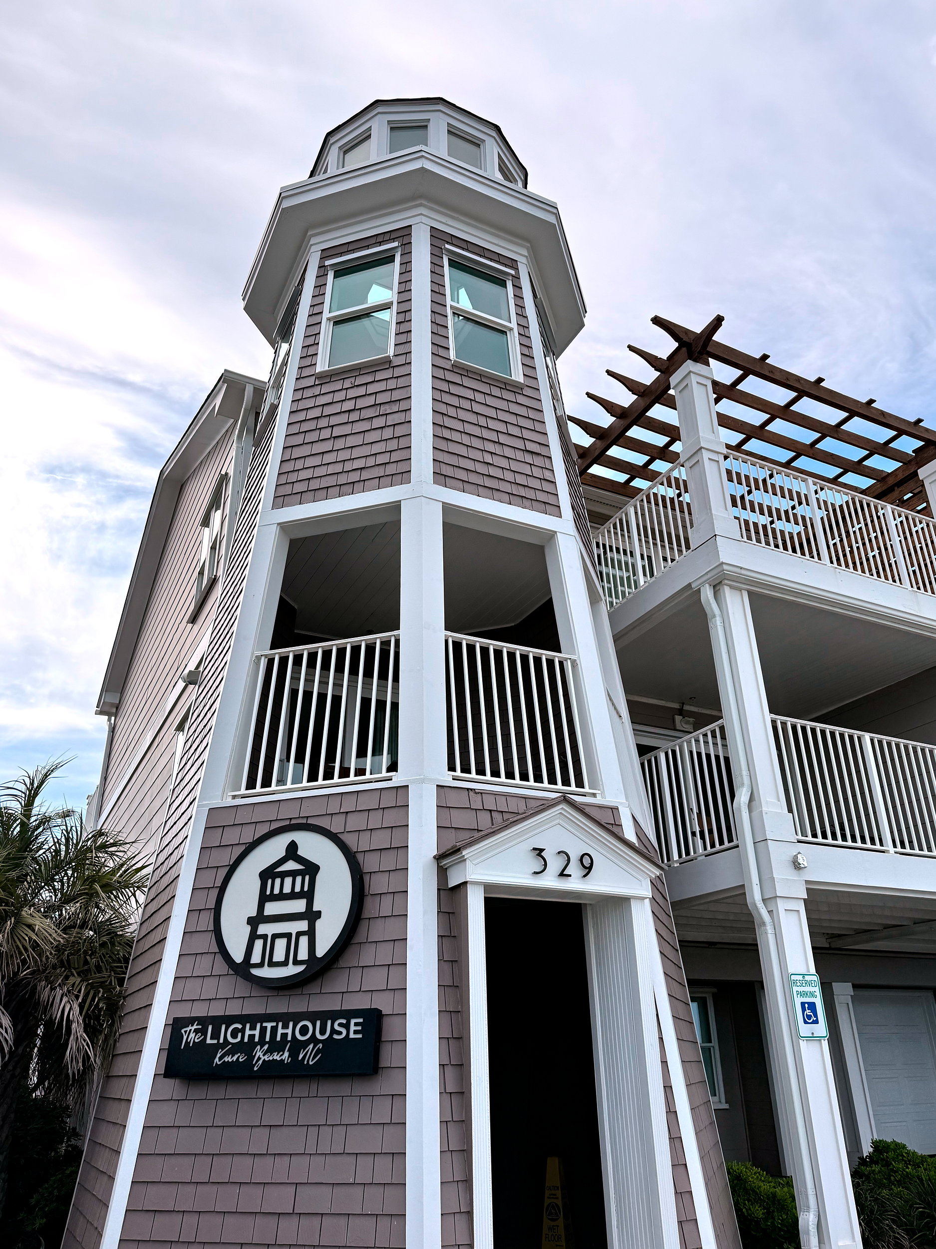 Exterior of The Lighthouse Inn in Kure Beach, North Carolina, featuring a lighthouse-style tower with signage and multiple balconies, and a clear blue sky.